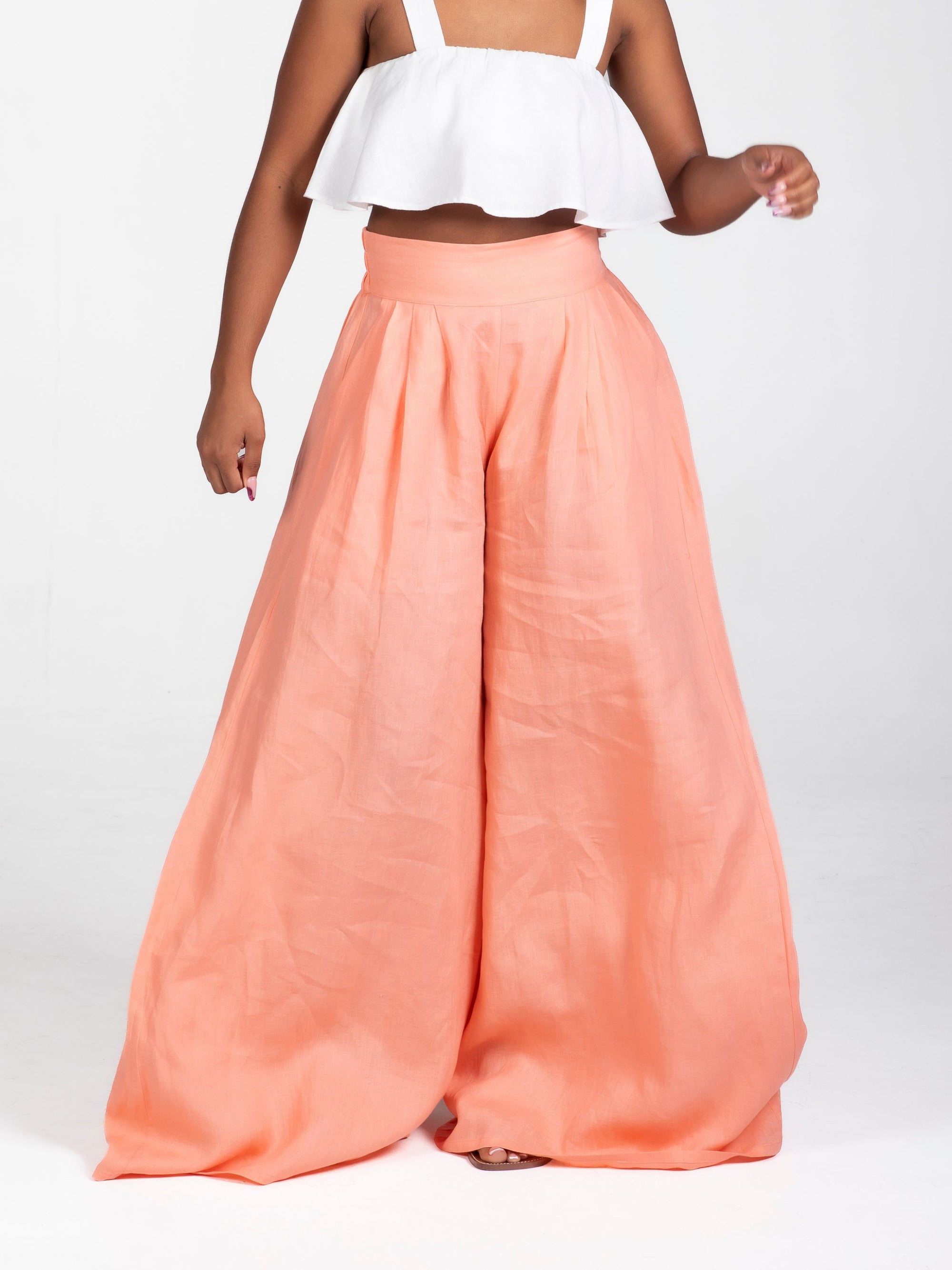 Model wearing high-waisted, wide-leg linen pants in a soft peach tone, styled with a white cropped ruffle top. The trousers feature a relaxed fit and deep side seam pockets, offering an easy, breezy silhouette for warm weather.