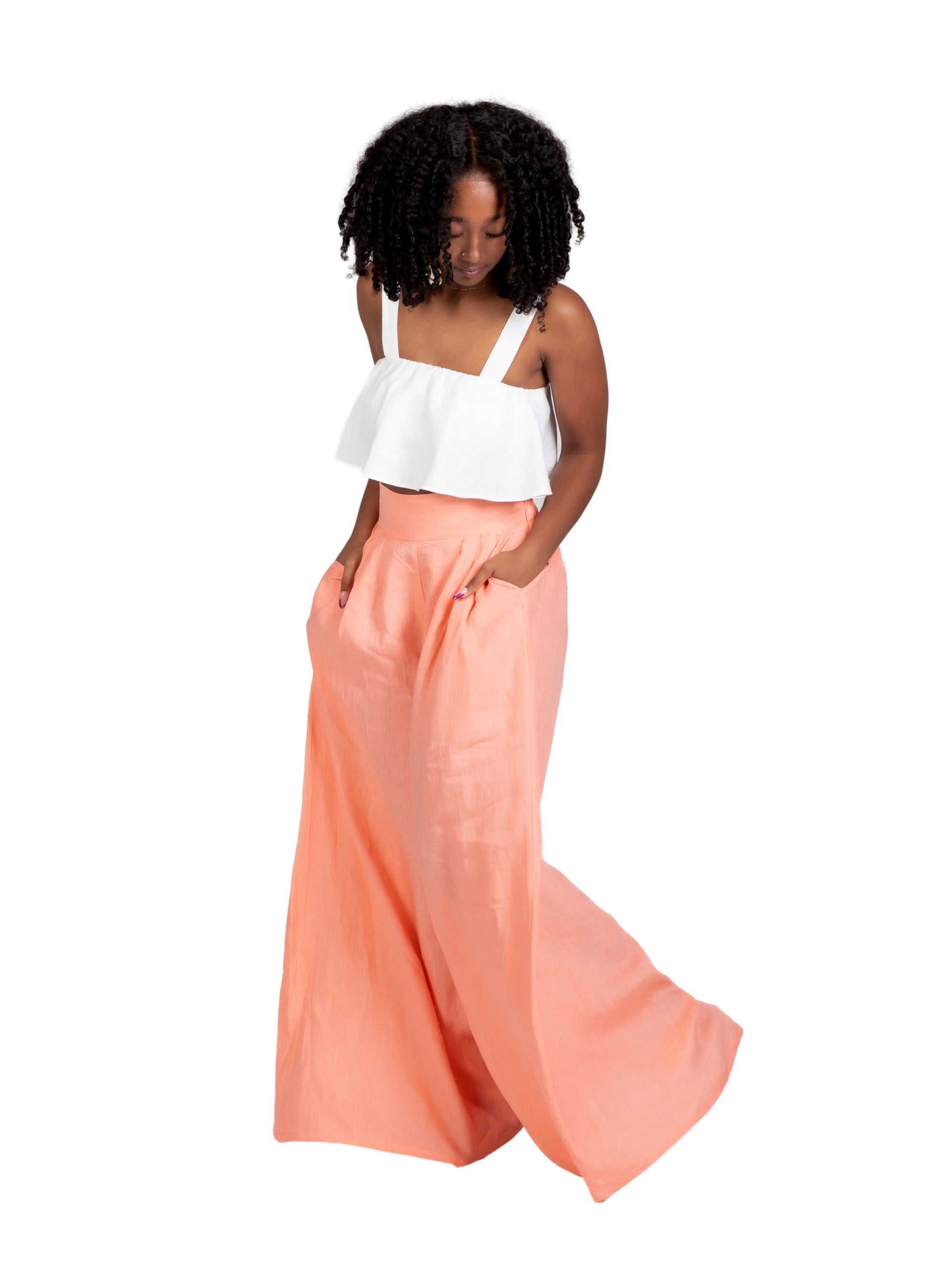 Model wearing high-waisted, wide-leg linen pants in a soft peach tone, styled with a white cropped ruffle top. The trousers feature a relaxed fit and deep side seam pockets, offering an easy, breezy silhouette for warm weather.