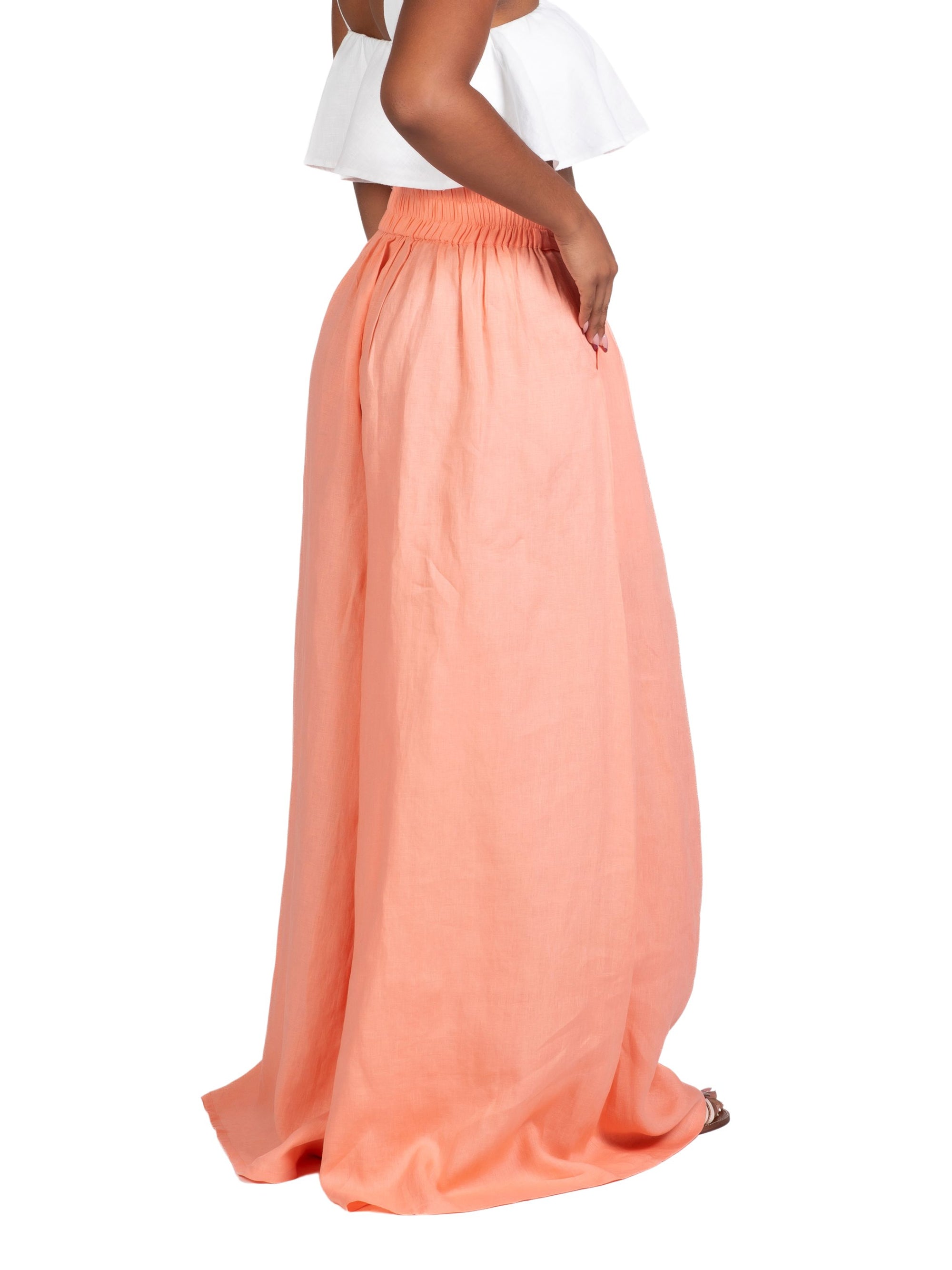 Model wearing high-waisted, wide-leg linen pants in a soft peach tone, styled with a white cropped ruffle top. The trousers feature a relaxed fit and deep side seam pockets, offering an easy, breezy silhouette for warm weather.