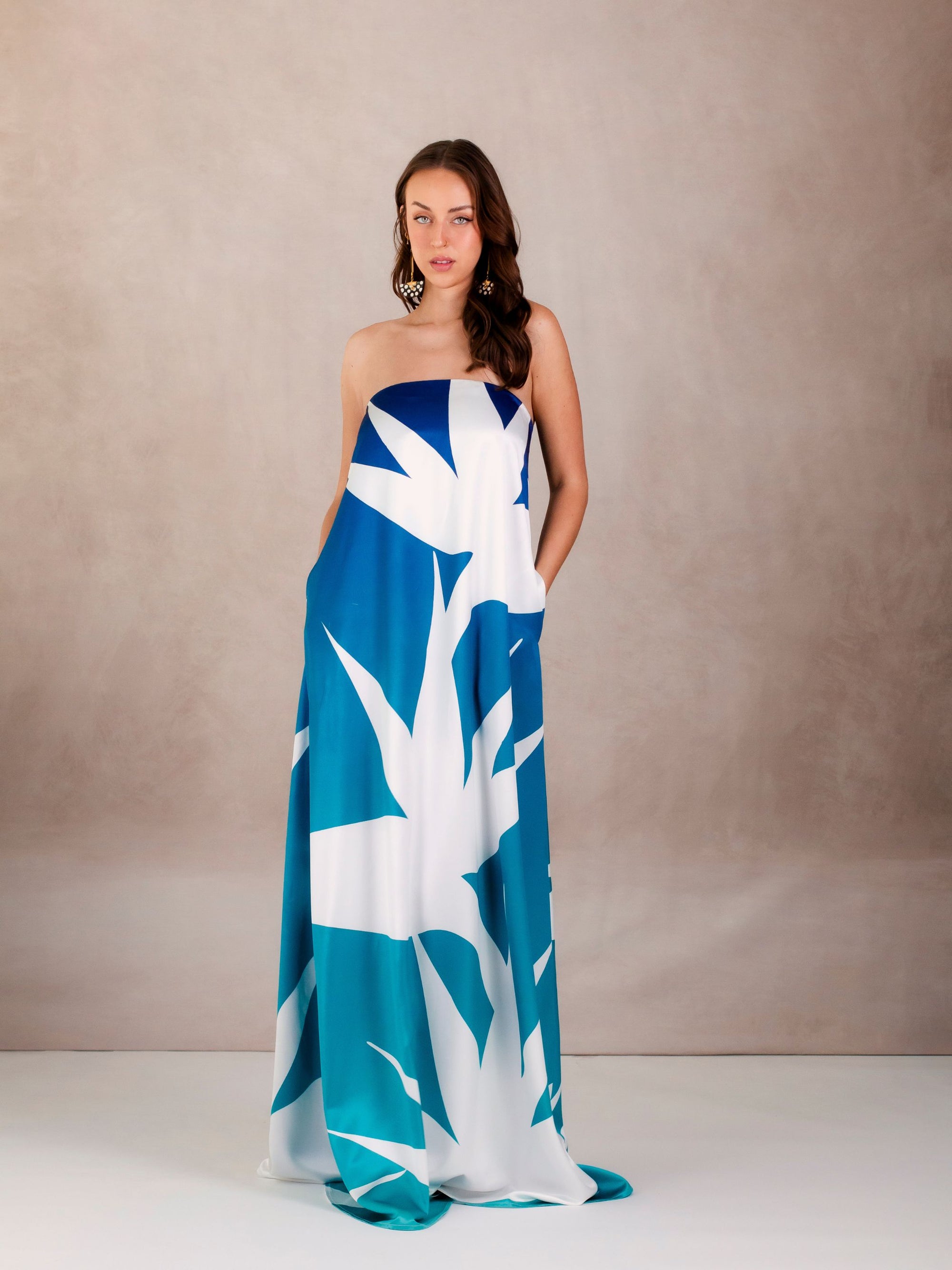A woman modeling a strapless, blue and white tropical print gown with side seam pockets. The dress drapes gracefully to the floor, highlighting its elegant, flowing silhouette.