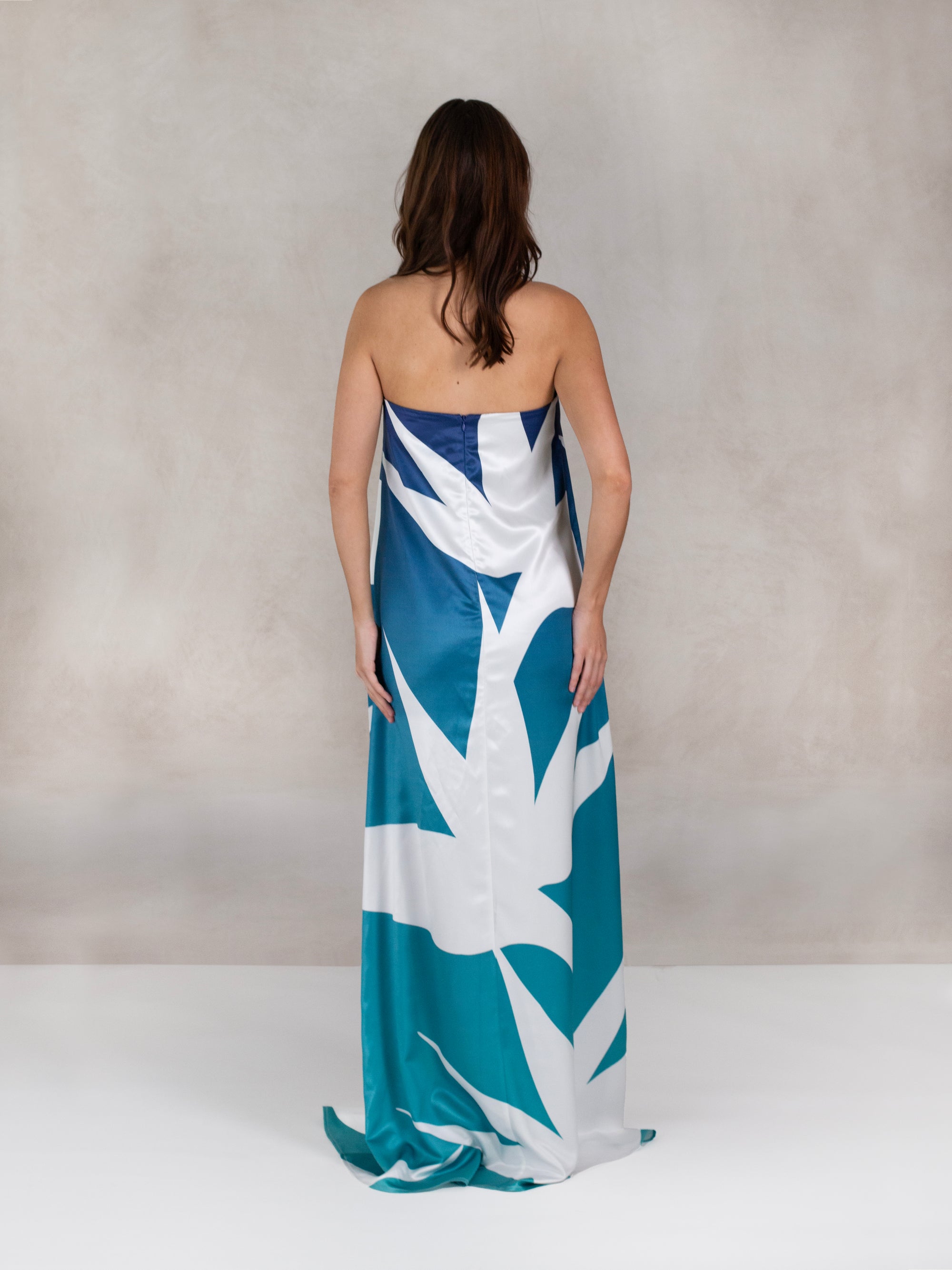 A woman wearing a strapless, floor-length gown featuring a bold, oversized tropical print in blue and white. The gown has a slight train and a fitted back with a concealed zipper.
