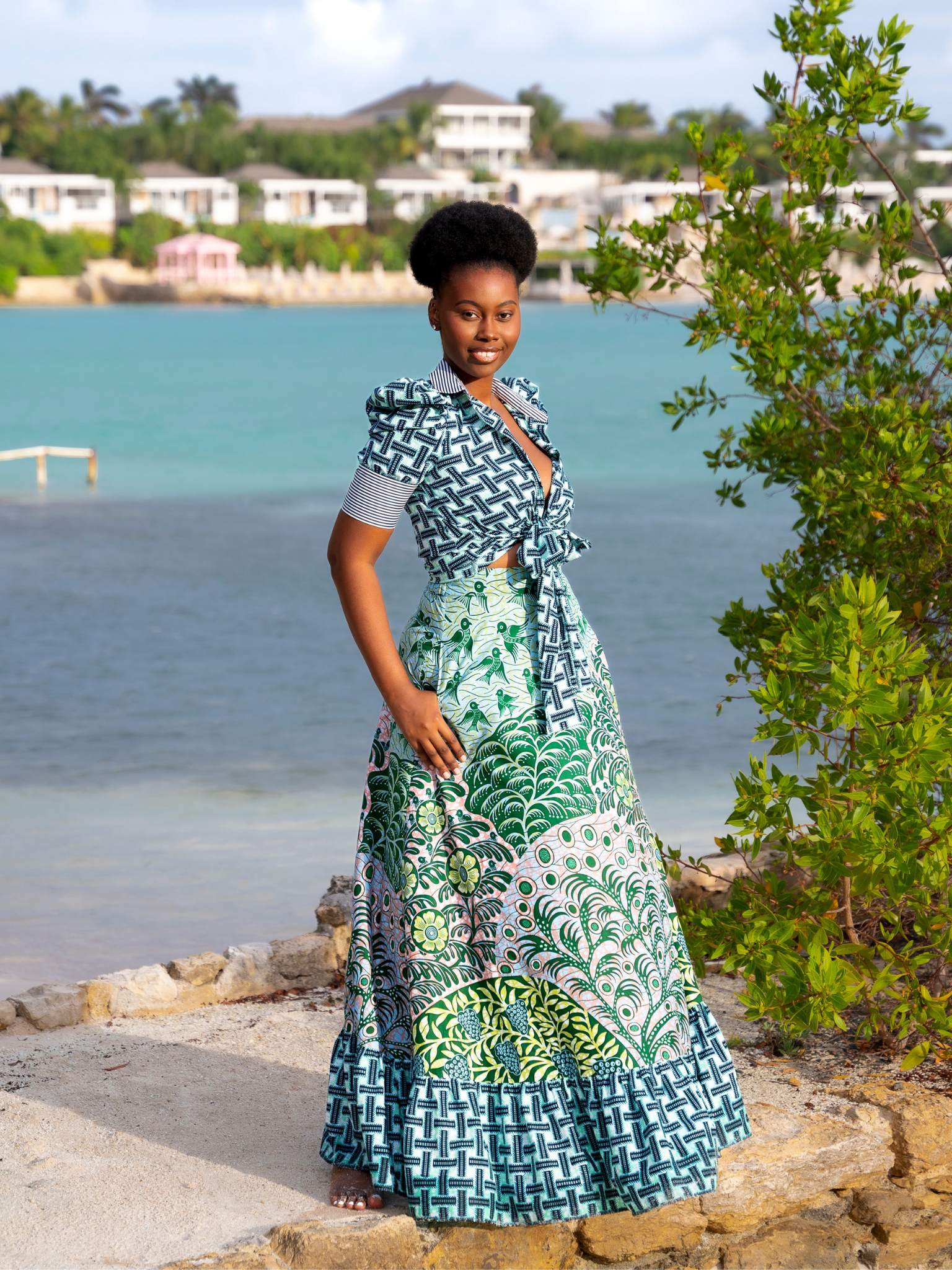 A woman stands on a stone path by turquoise waters, wearing the Chen Burkett New York Selah Top and Eden Maxi Skirt in a vibrant green and blue African print. The top features puffed sleeves and a tie-front, paired with a full-length skirt with tiered hem.