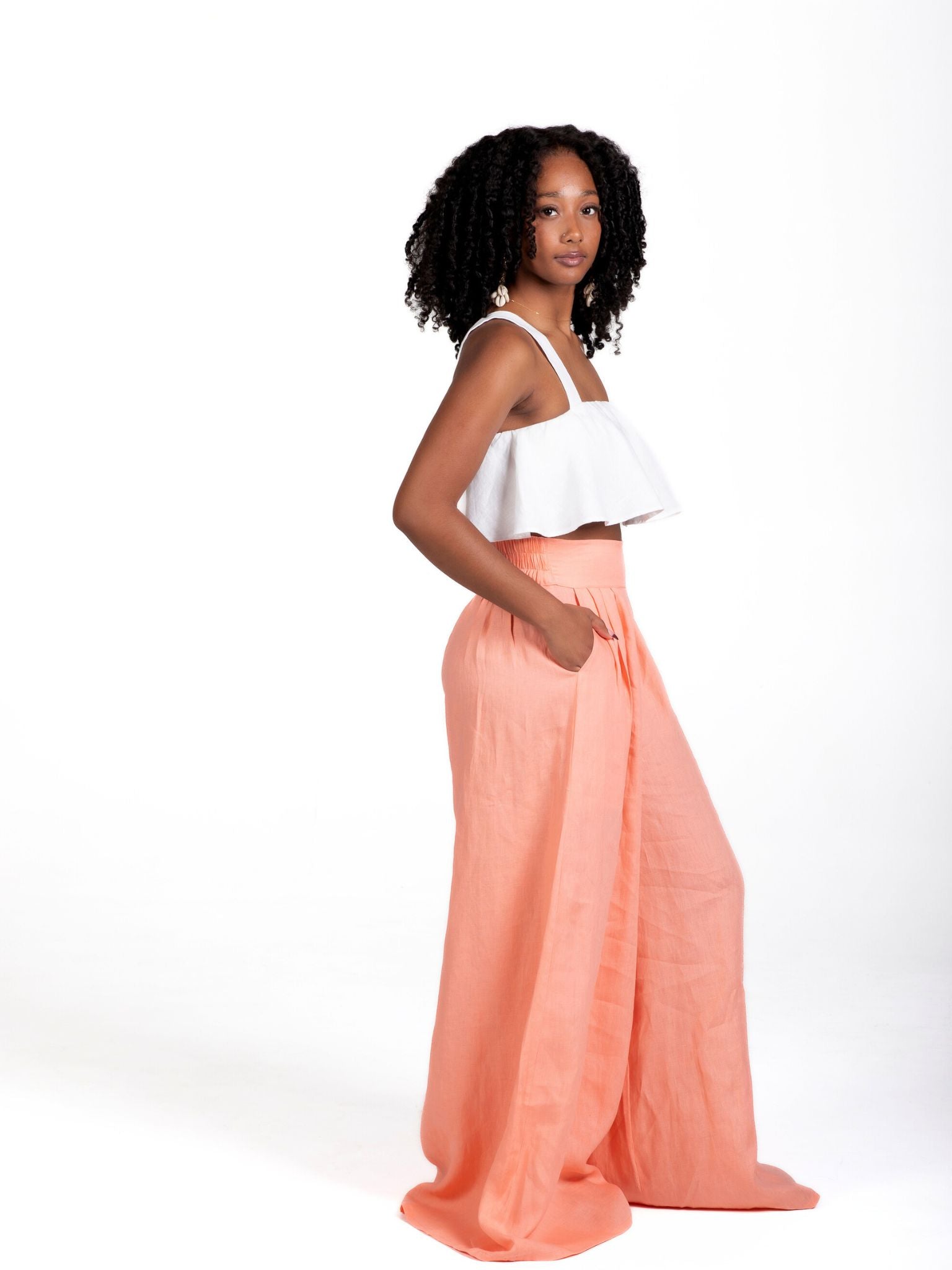 Model wearing high-waisted, wide-leg linen pants in a soft peach tone, styled with a white cropped ruffle top. The trousers feature a relaxed fit and deep side seam pockets, offering an easy, breezy silhouette for warm weather.