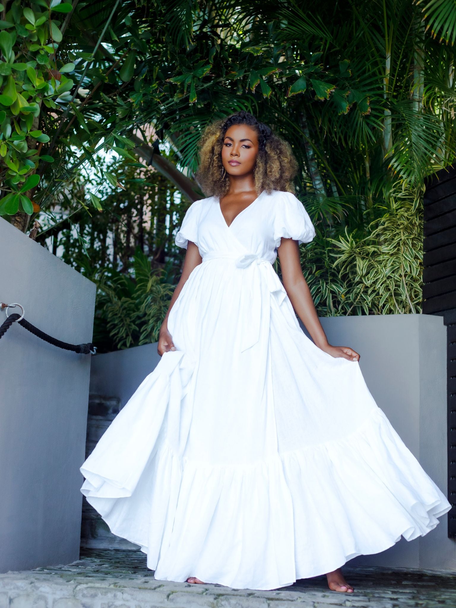 A tall woman in a white linen wrap dress stands on stone steps surrounded by lush greenery. The dress features puff sleeves, a tiered skirt, and a waist tie.