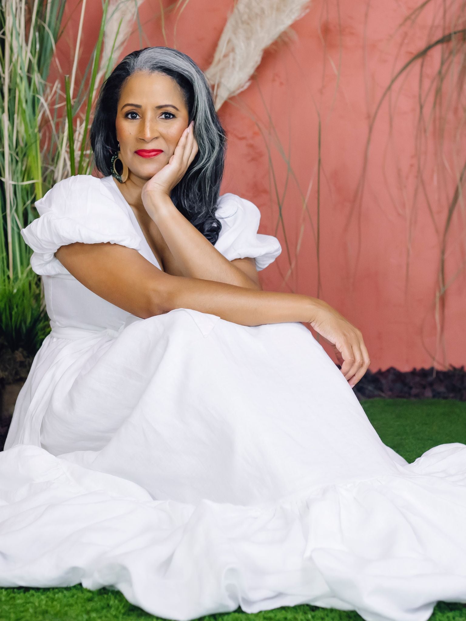 A woman with long salt-and-pepper hair wears a white linen wrap dress with puff sleeves and a tiered skirt. She is smiling and standing on green grass in front of a terracotta-colored wall surrounded by pampas grass and greenery.