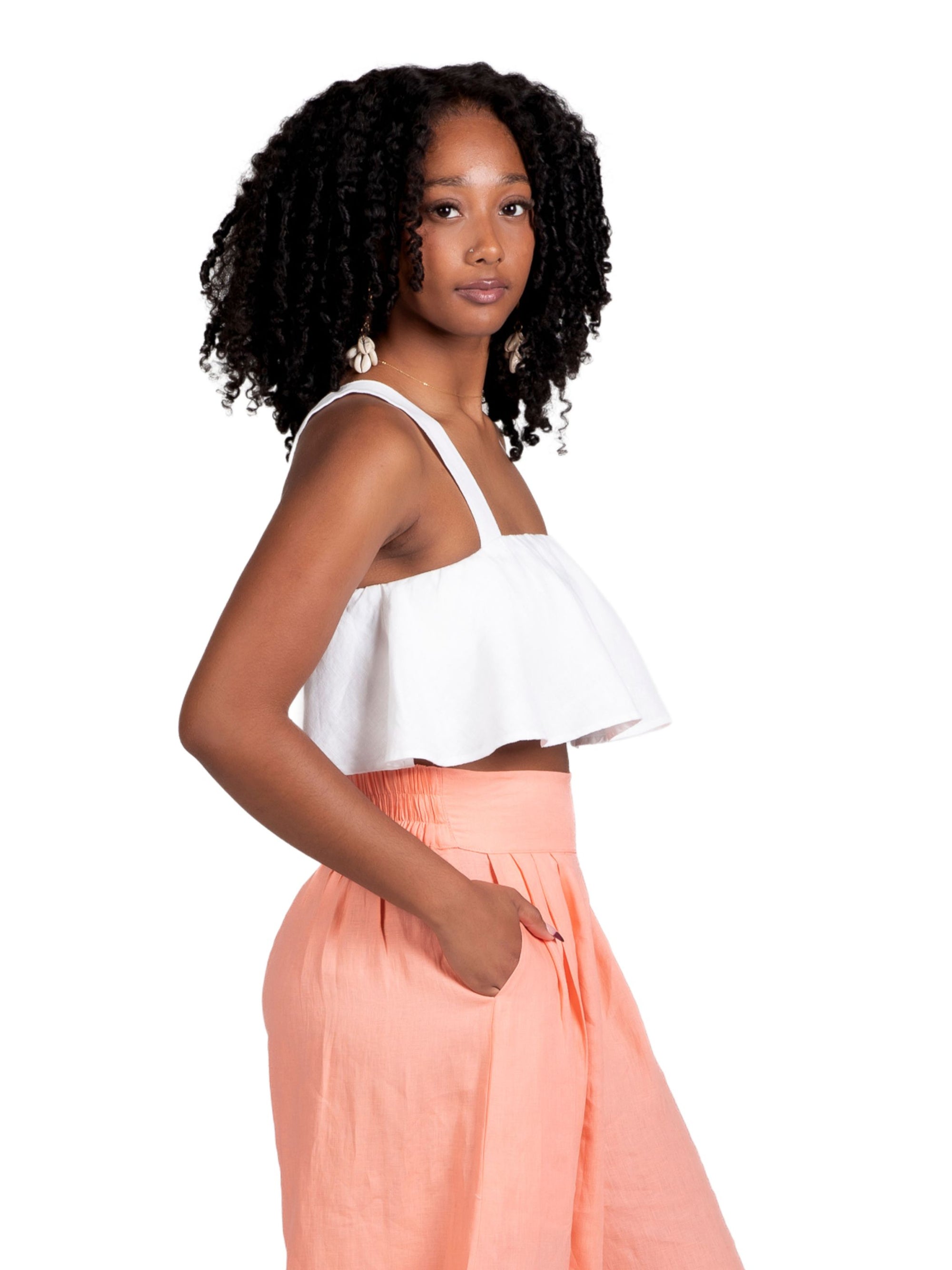 A young woman with natural curly hair wears the white Olii Linen Top paired with turquoise printed pants. She is smiling brightly with one hand in her pocket, posing against a white studio background.