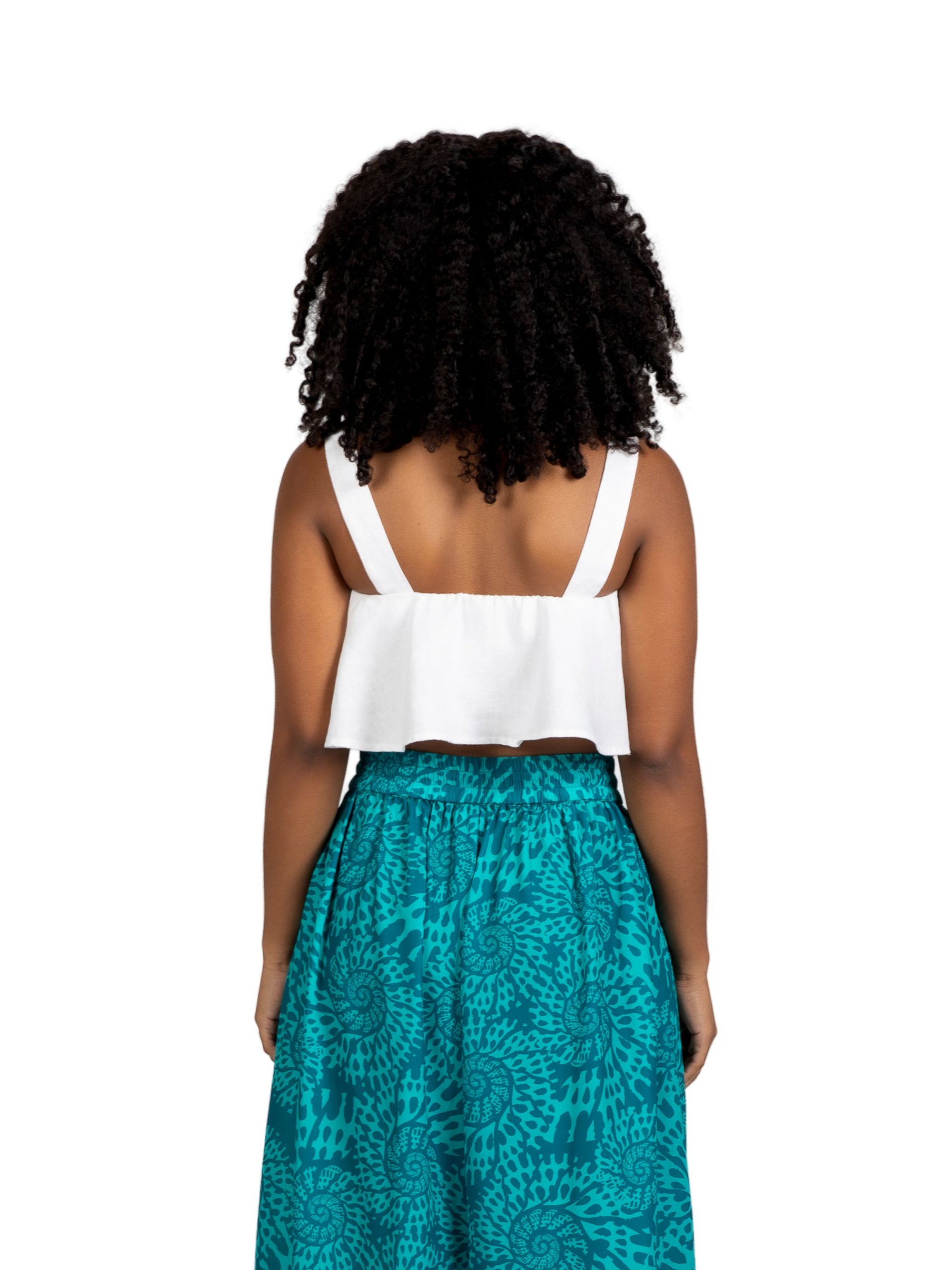 A young woman with natural curly hair wears the white Olii Linen Top paired with turquoise printed pants. She is smiling brightly with one hand in her pocket, posing against a white studio background.