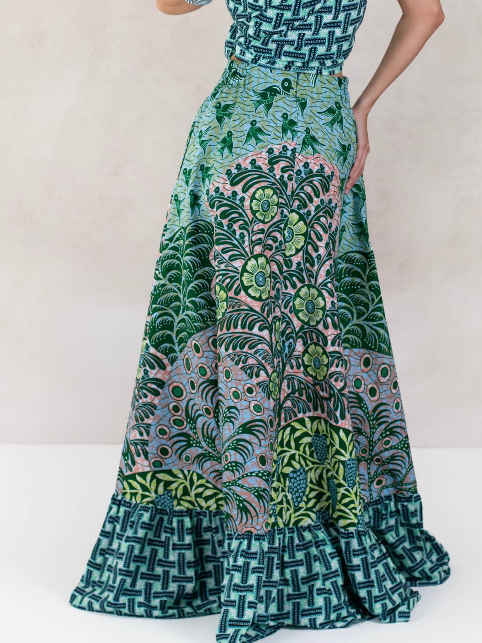 Model in studio wearing the Selah Top and Eden Maxi Skirt by Chen Burkett New York. The look features bold mixed prints in shades of teal, green, and pink, with a structured short-sleeve blouse tied at the waist and a flowing, voluminous skirt with pockets.