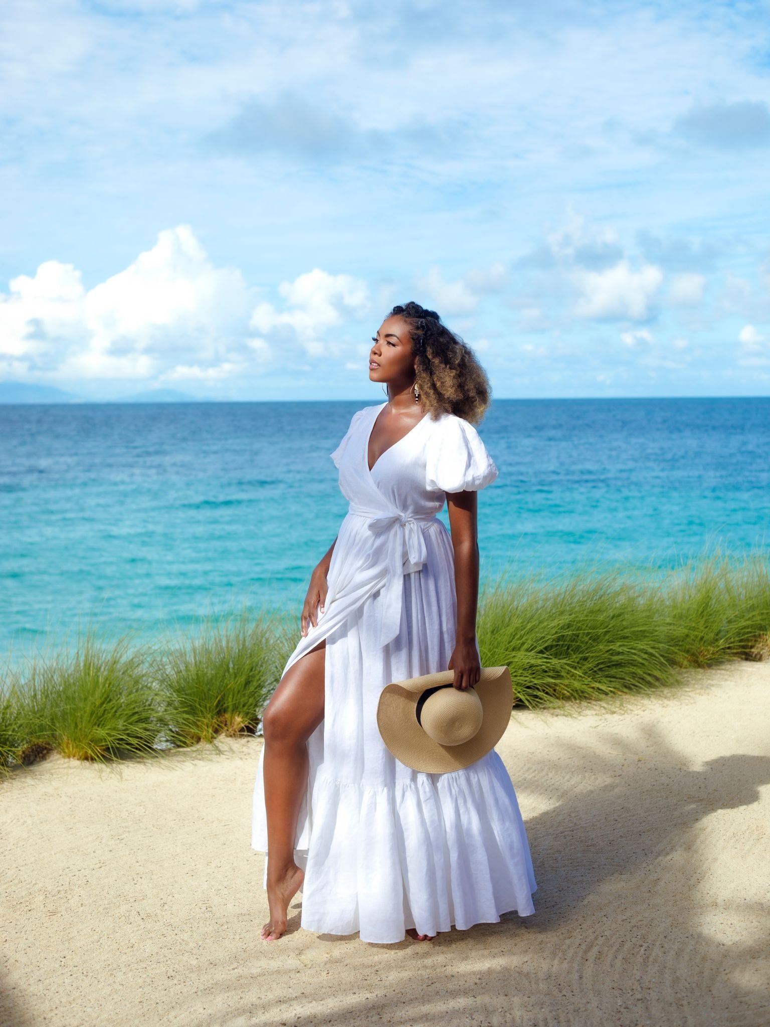 The model wears the Marigot Bay Linen Dress barefoot on a sandy beach with turquoise ocean in the background, holding a sunhat in one hand and gazing toward the sea.