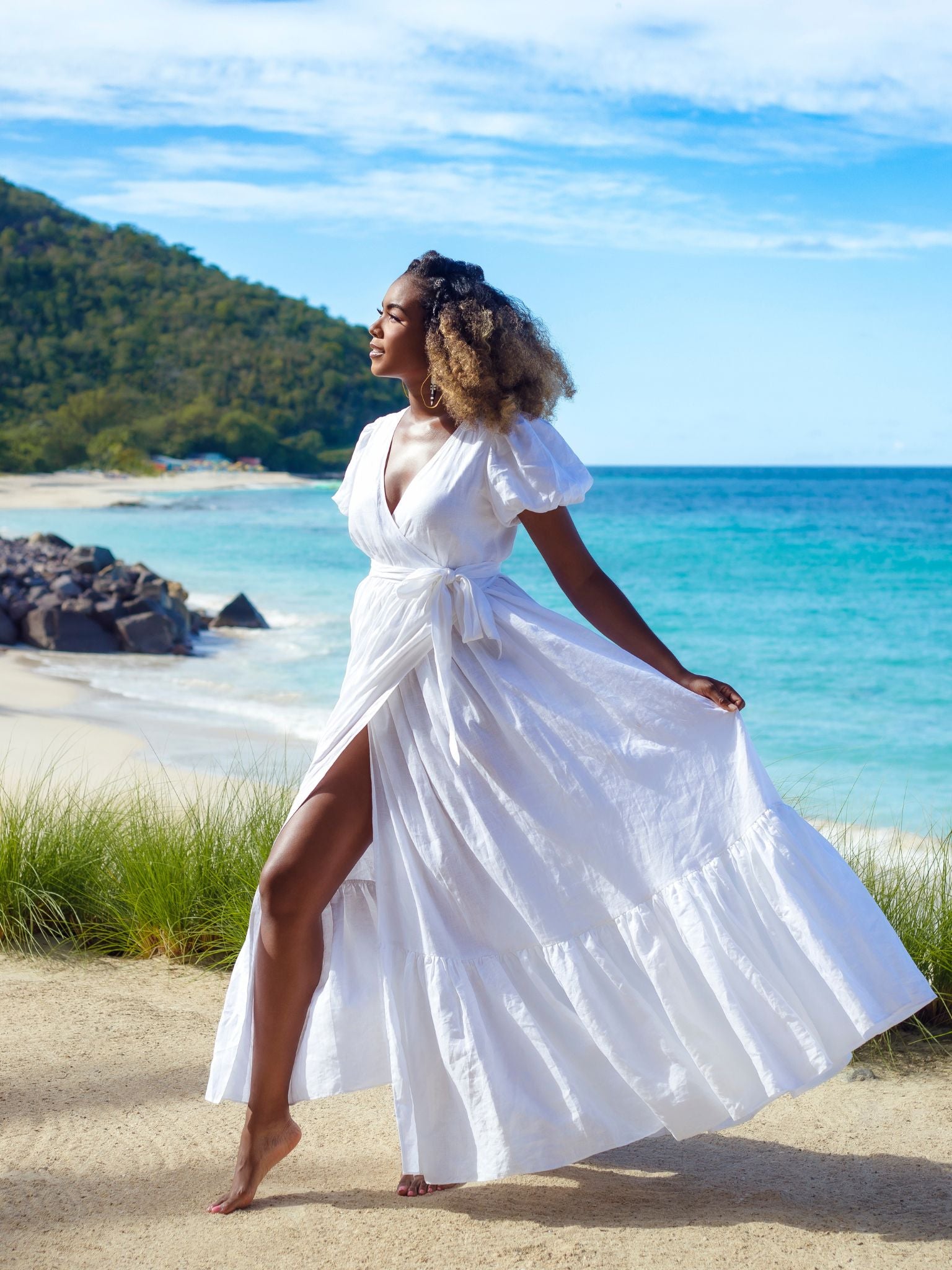 The model wears the Marigot Bay Linen Dress barefoot on a sandy beach with turquoise ocean in the background, holding a sunhat in one hand and gazing toward the sea.