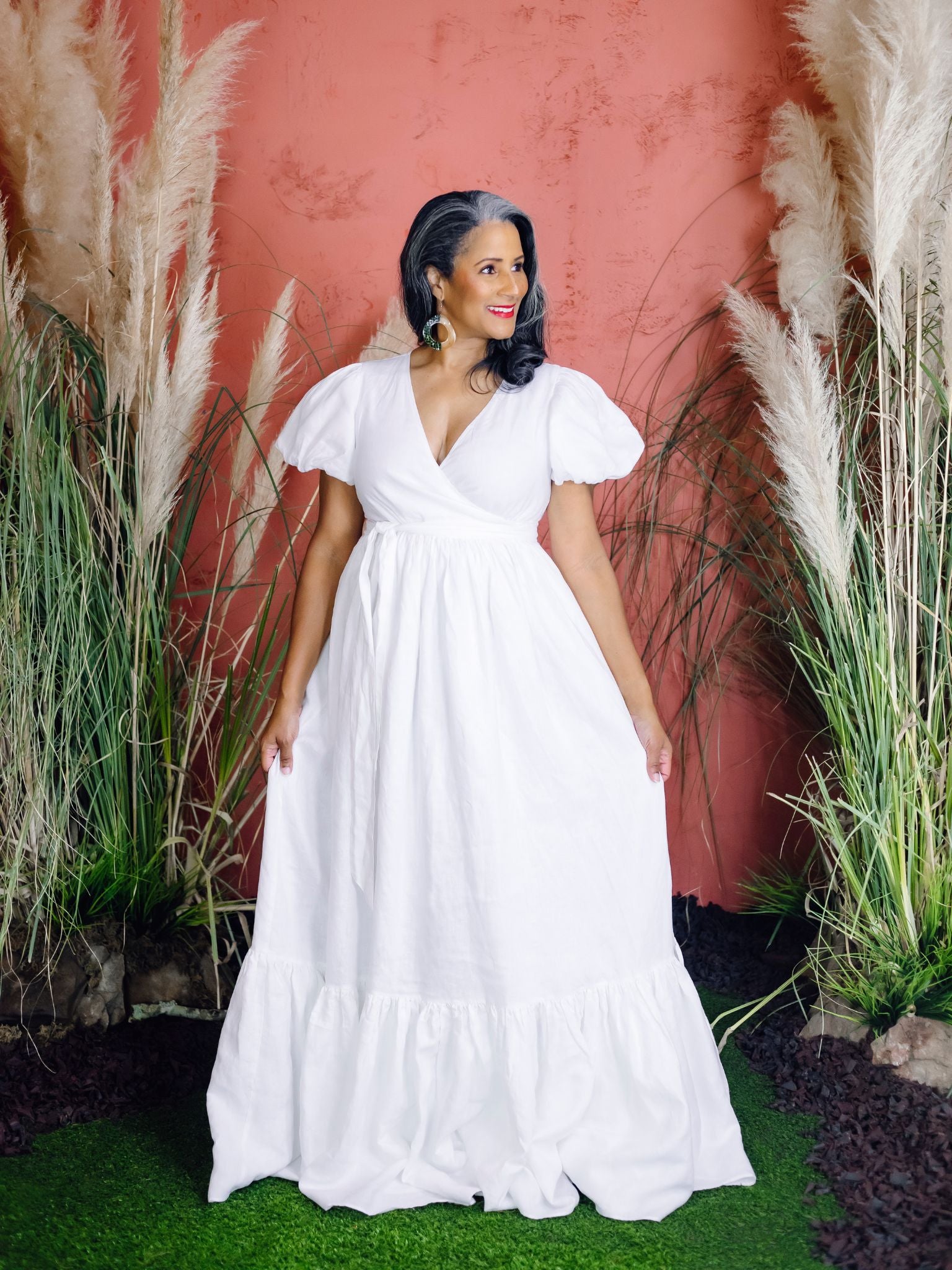 A woman with long salt-and-pepper hair wears a white linen wrap dress with puff sleeves and a tiered skirt. She is smiling and standing on green grass in front of a terracotta-colored wall surrounded by pampas grass and greenery.