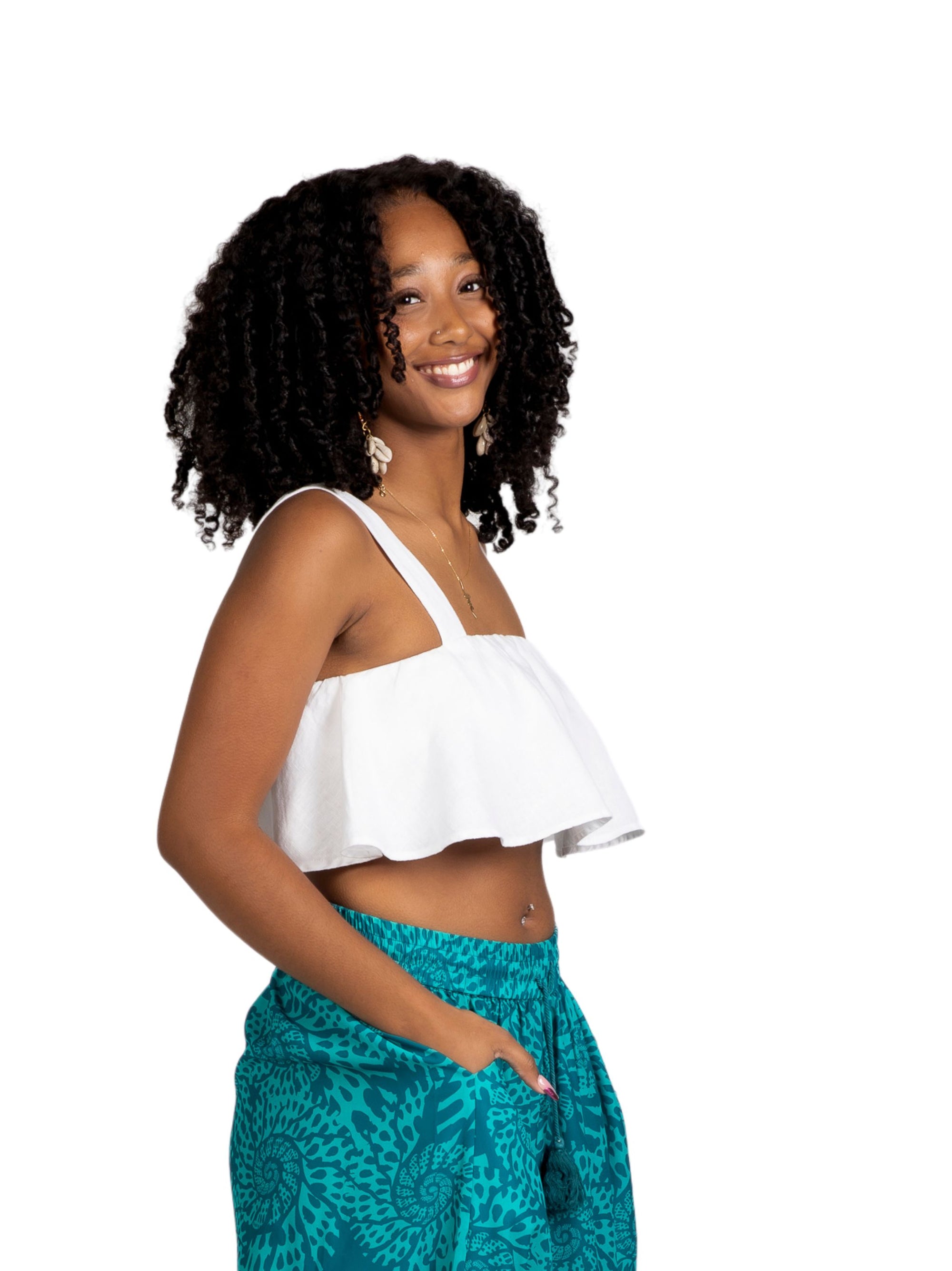 A young woman with natural curly hair wears the white Olii Linen Top paired with turquoise printed pants. She is smiling brightly with one hand in her pocket, posing against a white studio background.