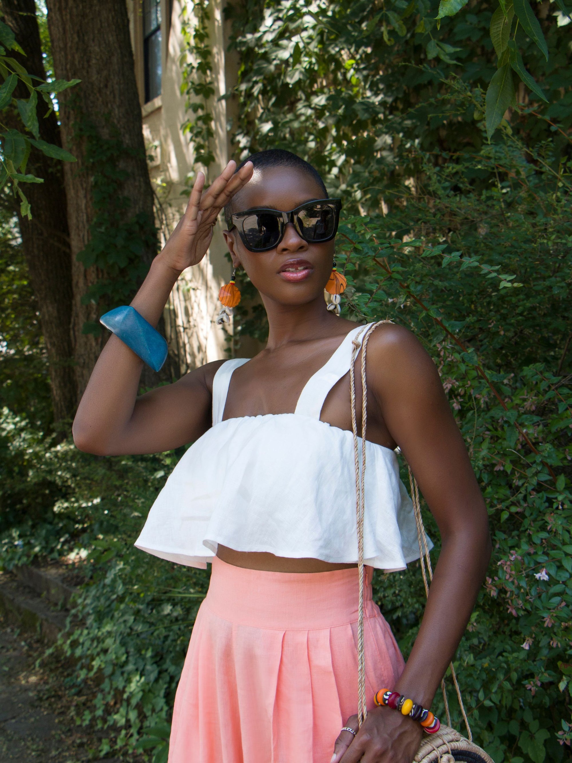 A young woman with low haircut and sunglasses wears the white Olii Linen Top paired with turquoise printed pants. She is smiling brightly with one hand in her pocket, posing against a white studio background.