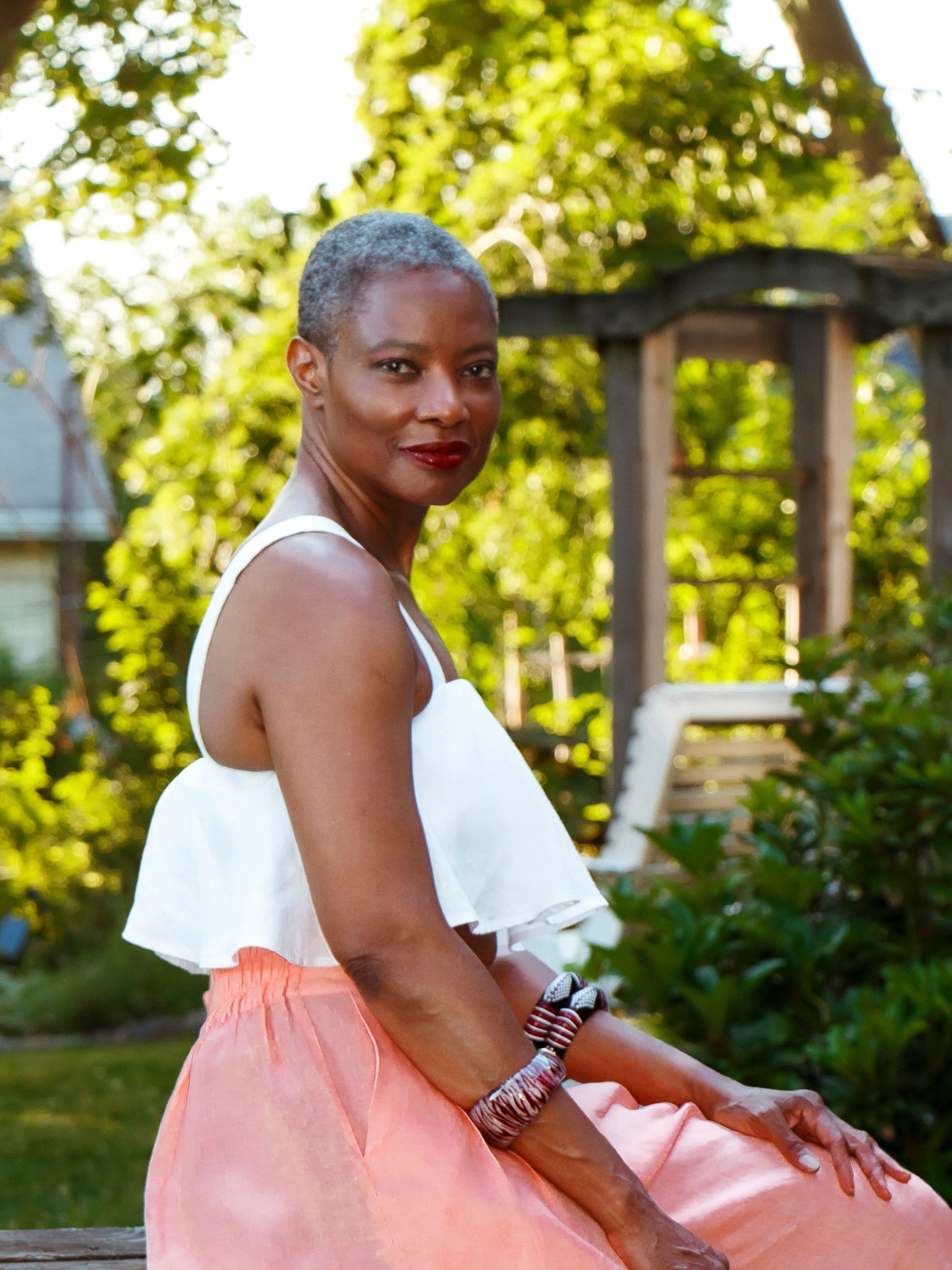 A mature woman with short grey hair sits outdoors wearing the Olii Linen Top and peach-colored Paradise Pants, accessorized with bold bangles and red lipstick.