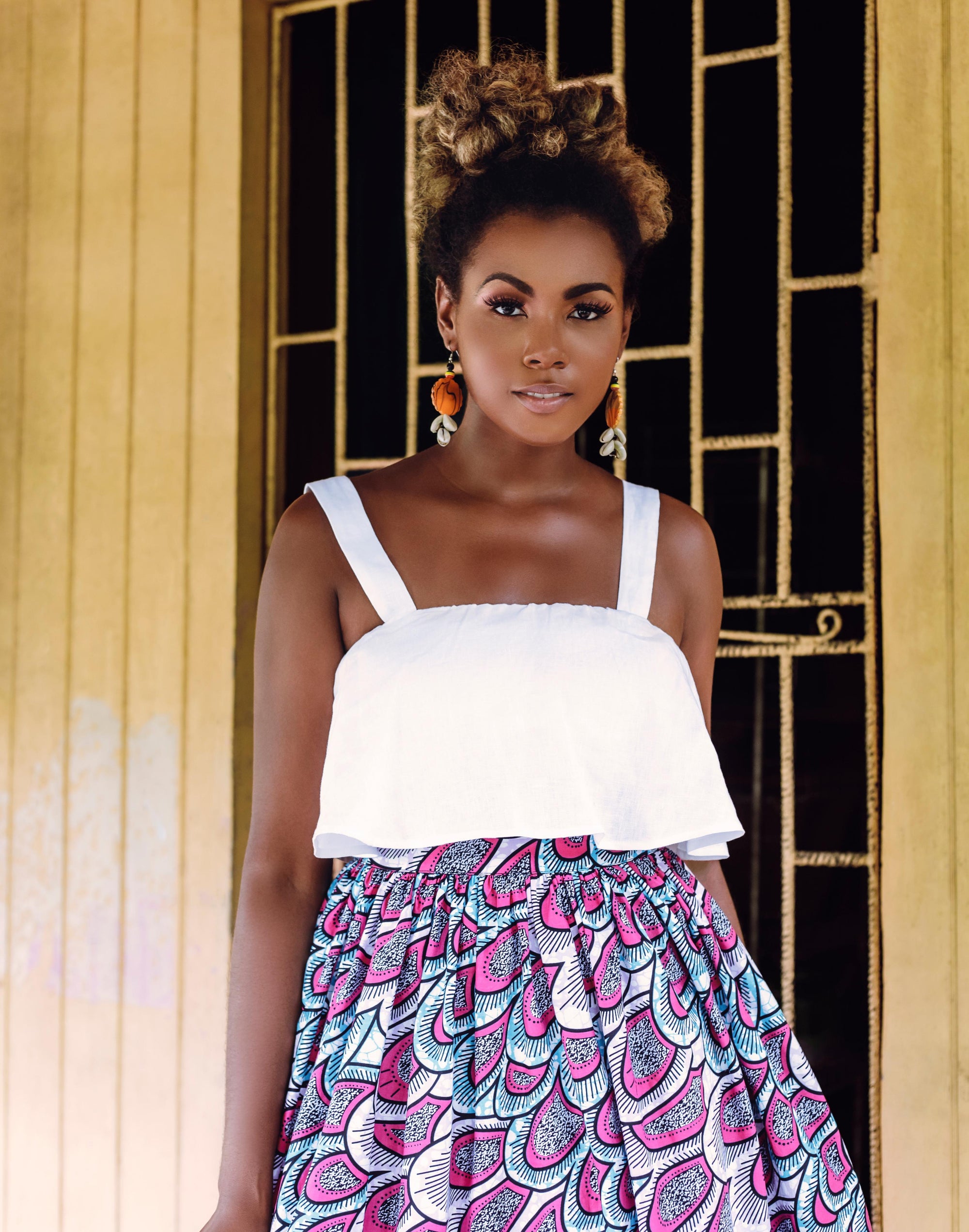 A woman with styled curls wears the white Olii Linen Top tucked into a colorful, pink and blue printed high-waisted skirt. She stands confidently in front of a vintage yellow doorway.