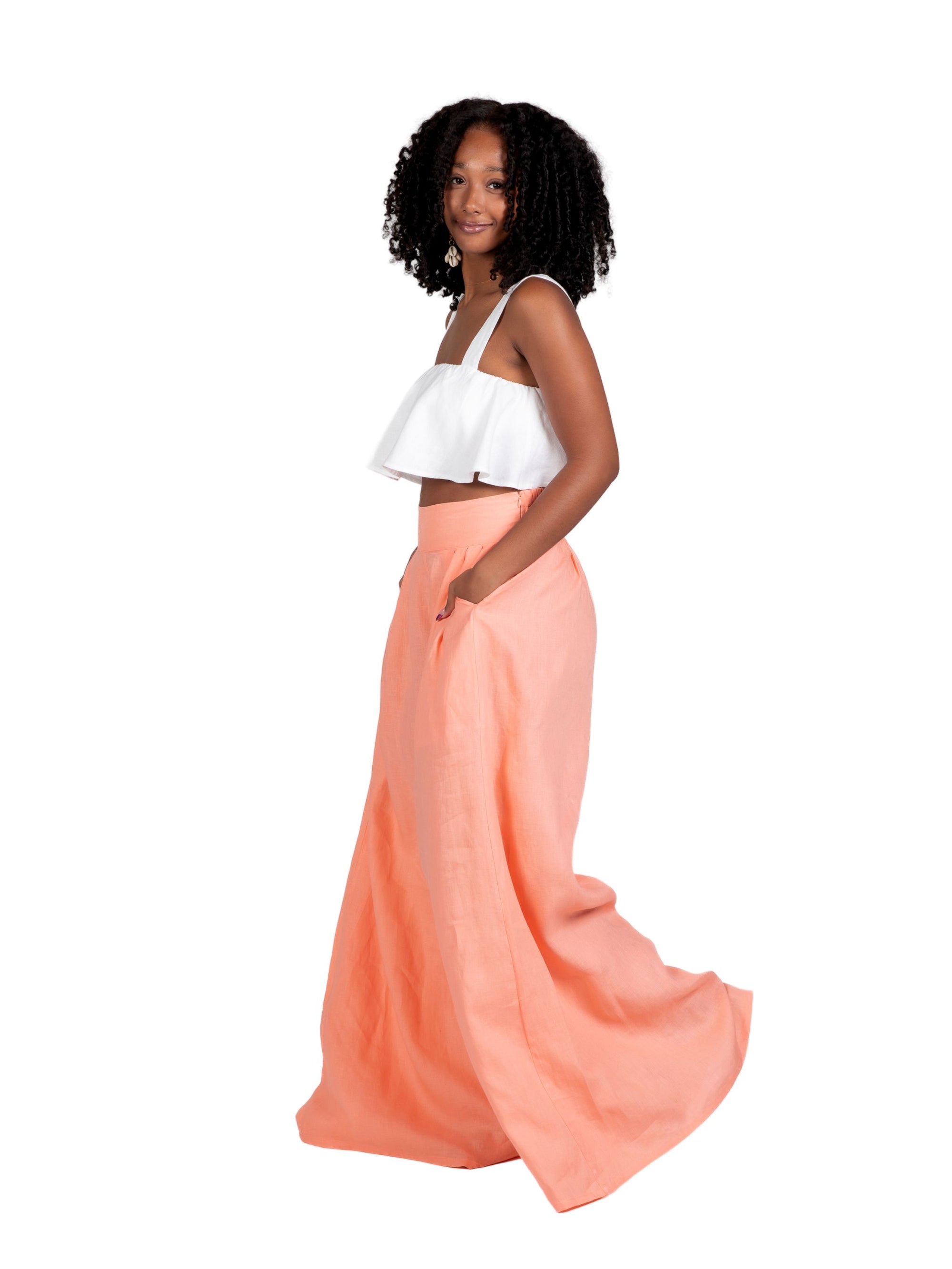 Model wearing high-waisted, wide-leg linen pants in a soft peach tone, styled with a white cropped ruffle top. The trousers feature a relaxed fit and deep side seam pockets, offering an easy, breezy silhouette for warm weather.