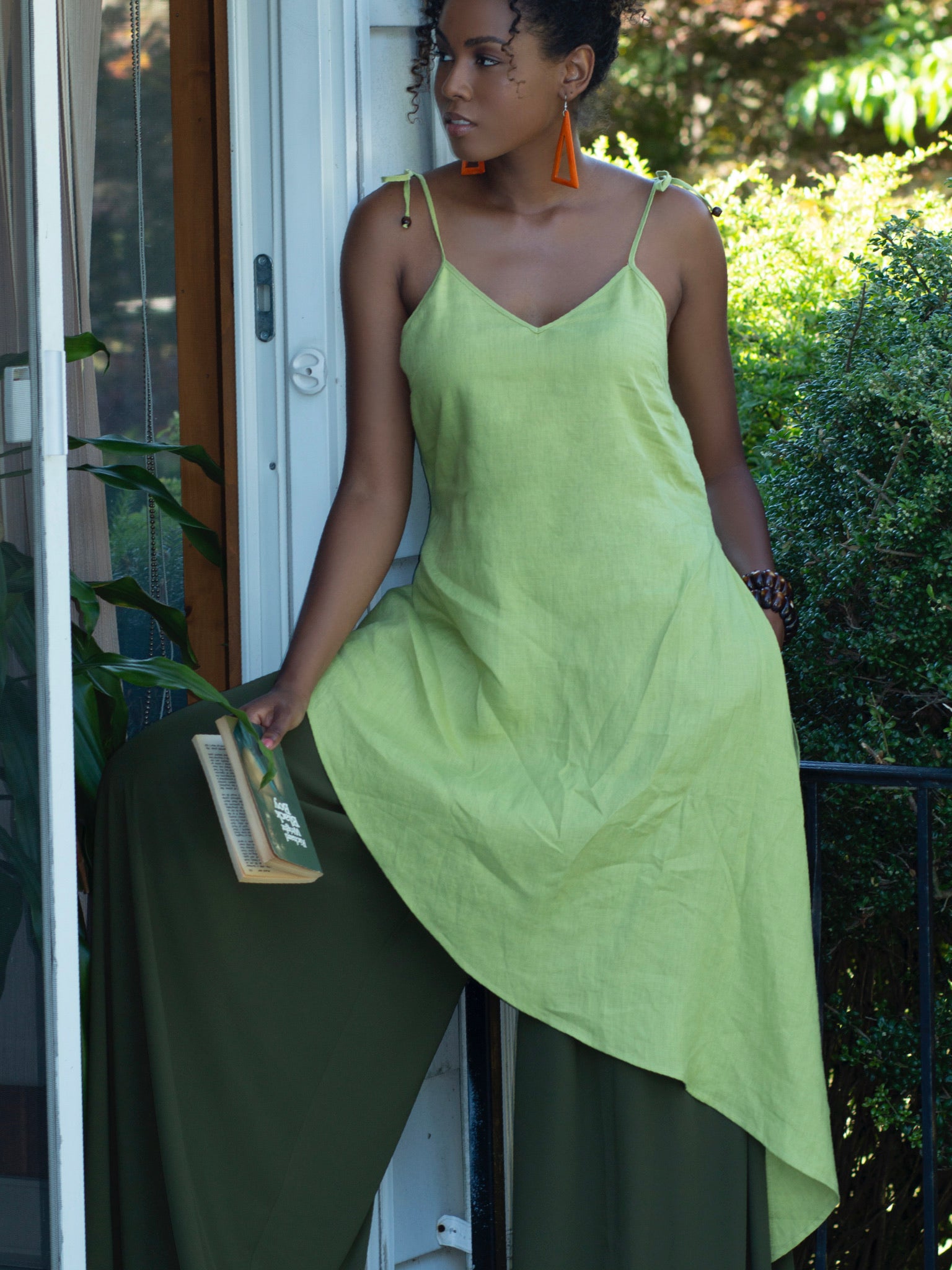 Black model standing in doorway holding a book to her side, wearing lime green linen tunic with spaghetti straps, gentle v-shaped neckline and long asymmetric hemline.