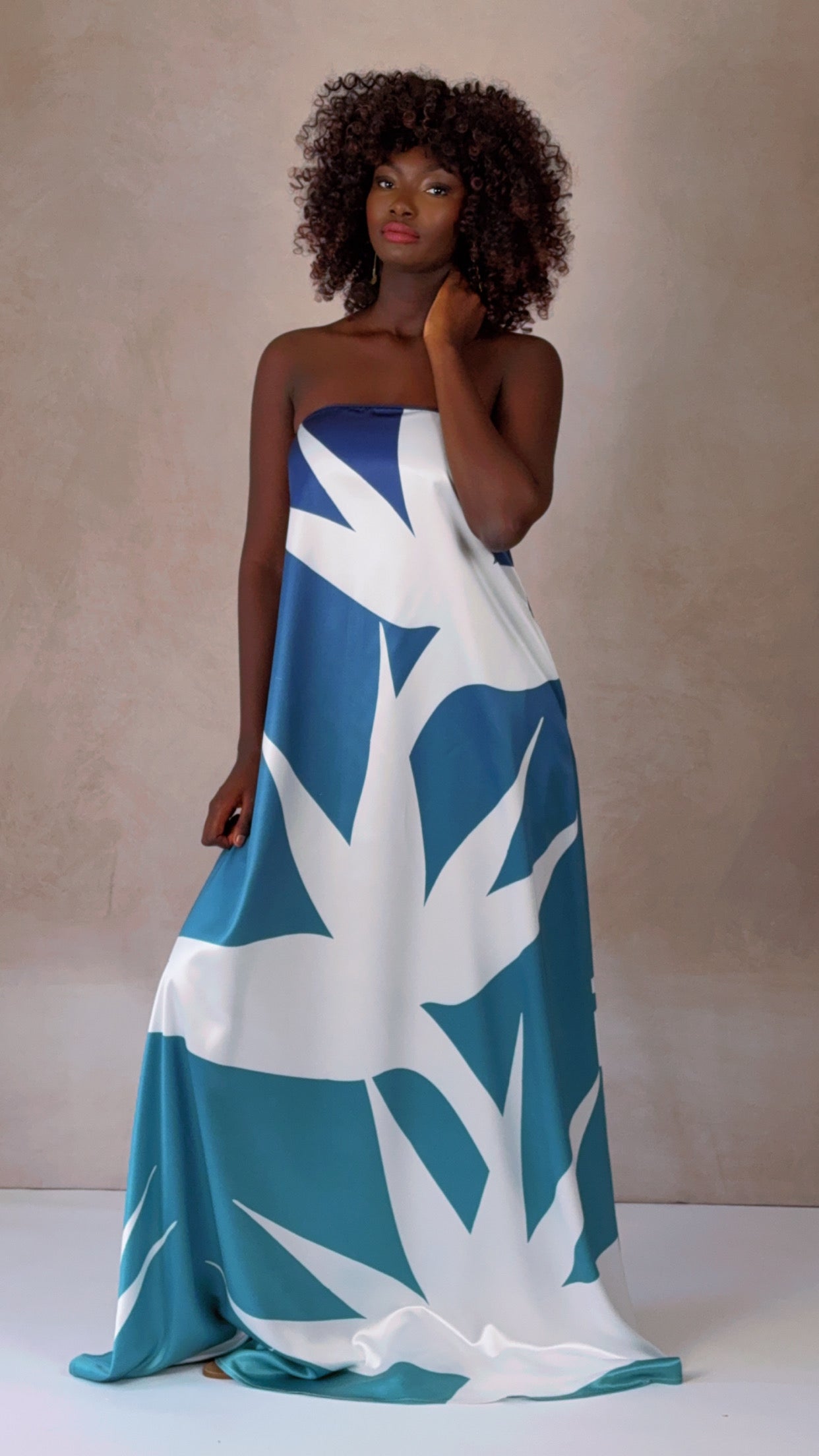 A woman modeling a strapless, blue and white tropical print gown with side seam pockets. The dress drapes gracefully to the floor, highlighting its elegant, flowing silhouette.