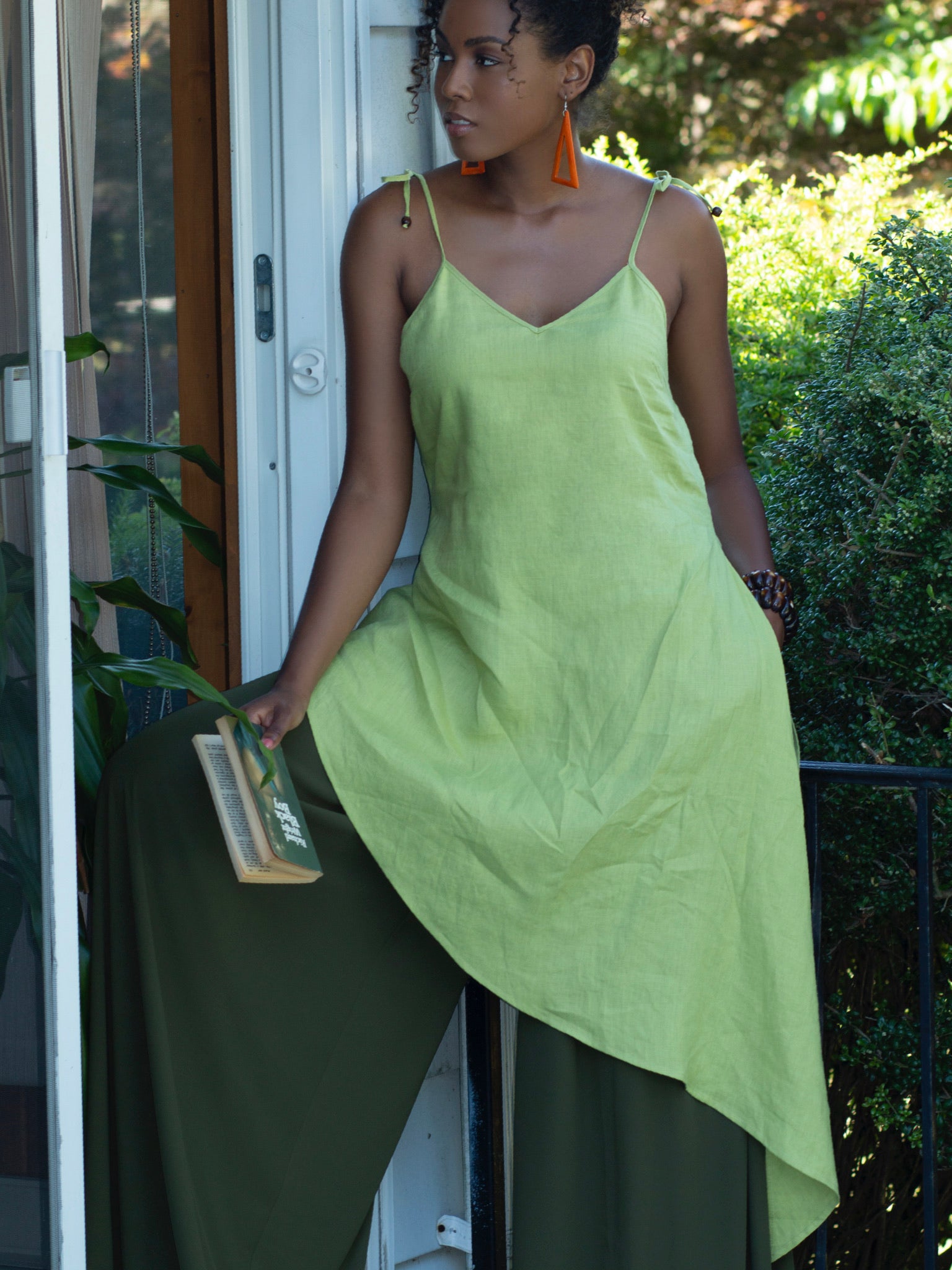 Black model standing in doorway holding a book to her side, wearing lime green  linen tunic with spaghetti straps, gentle v-shaped neckline and long asymmetric hemline. 
