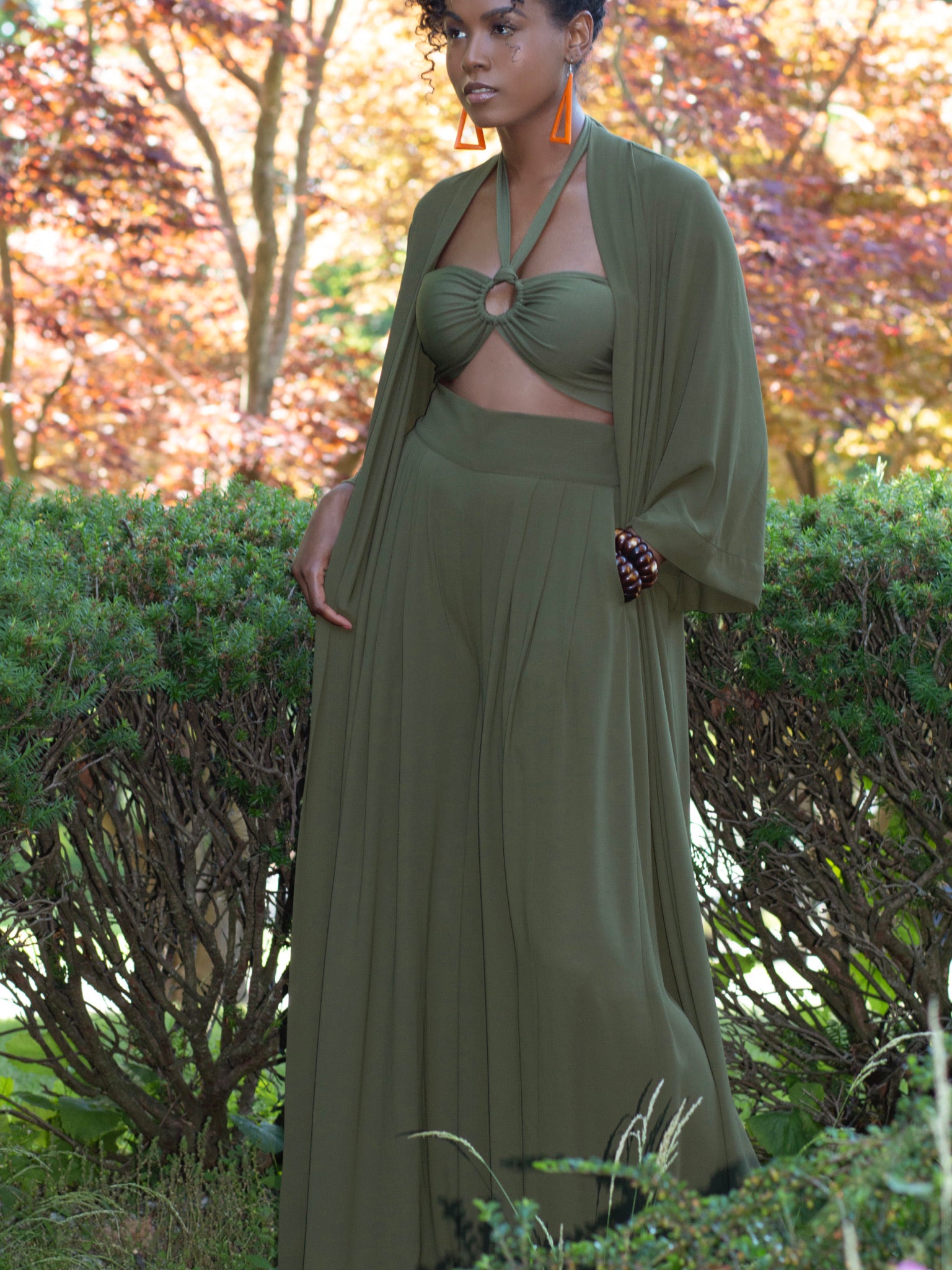 Black model standing with hand in pocket of dark green sheer fabric, wide legged trousers and matching dark olive green full length, open fronted duster. Duster has wide, loose sleeves