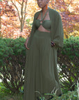 Black model standing with hand in pocket of dark green sheer fabric, wide legged trousers and matching dark olive green full length, open fronted duster. Duster has wide, loose sleeves